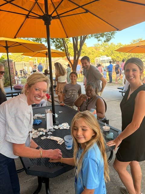MPA families catching up at outdoor picnic tables under umbrellas at Party on the Playground