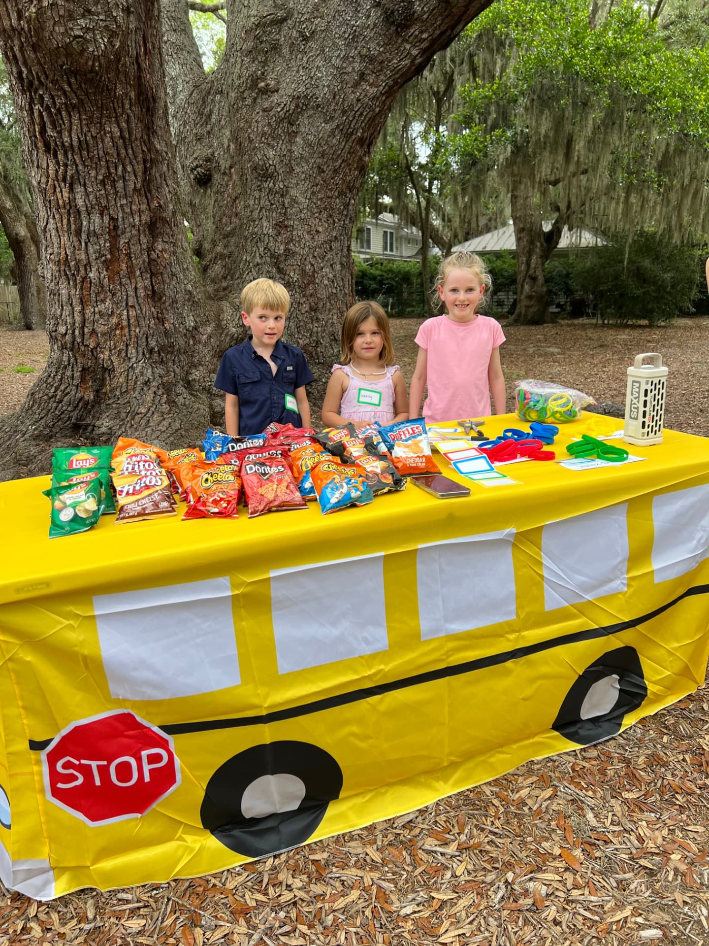 Young MPA students running a snack stand at a school bus themed table under a live oak